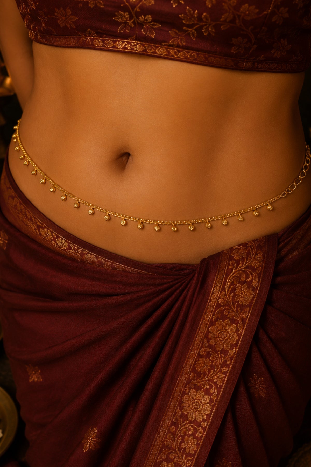 A close-up of a model's waist wearing a delicate gold chain belly drape (gharara) with small hanging bells, styled with a deep maroon and gold patterned sari.