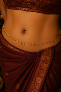 A close-up of a model's waist wearing a delicate gold chain belly drape (gharara) with small hanging bells, styled with a deep maroon and gold patterned sari.