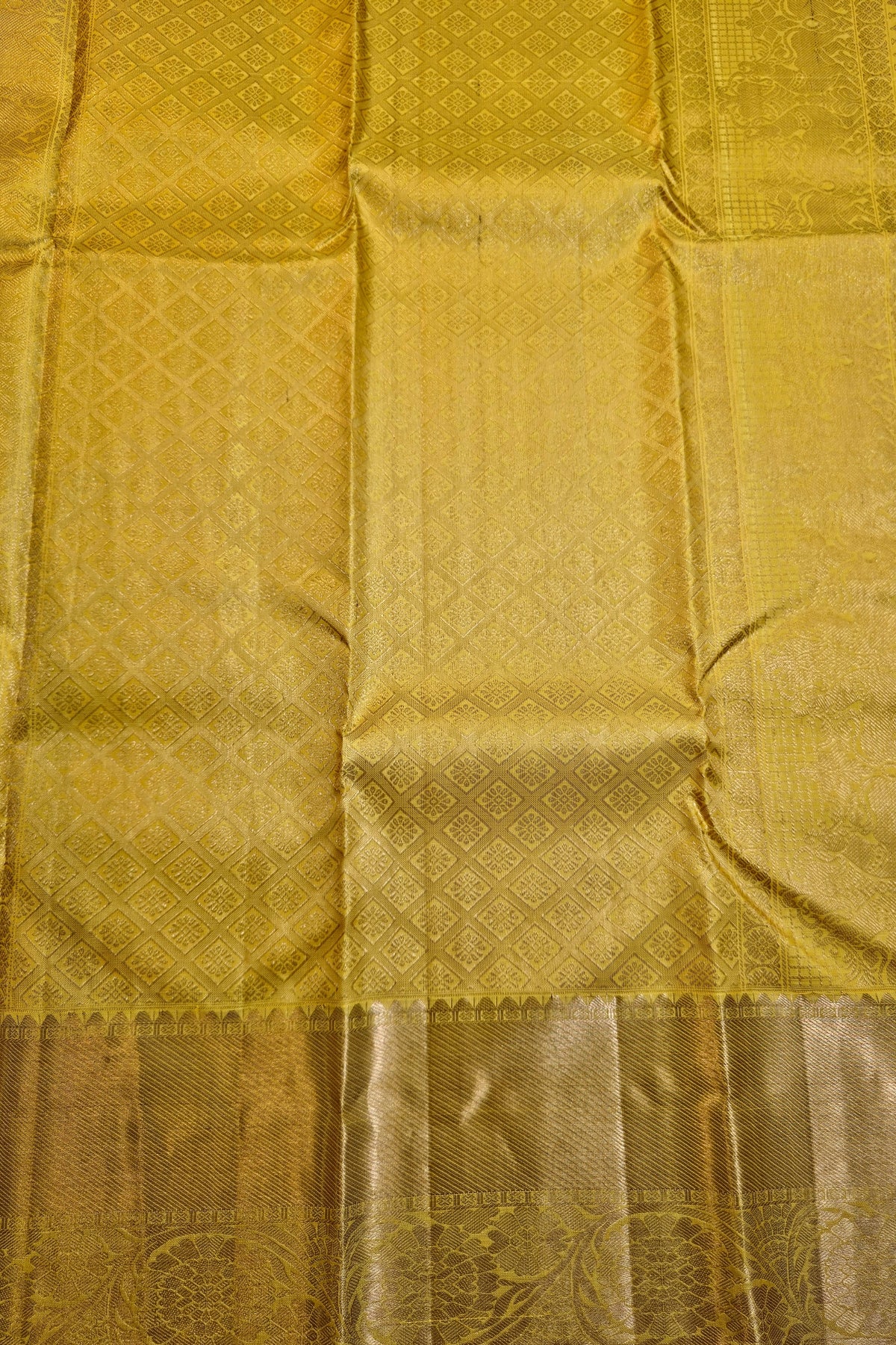 A close-up of a golden yellow silk fabric, likely a saree, showing its woven texture and a decorative border at the bottom.