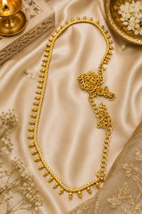 A gold-toned traditional necklace with a beaded upper section, chain lower section, and decorative clasp, displayed on draped satin fabric with small white flowers and ornamental props.