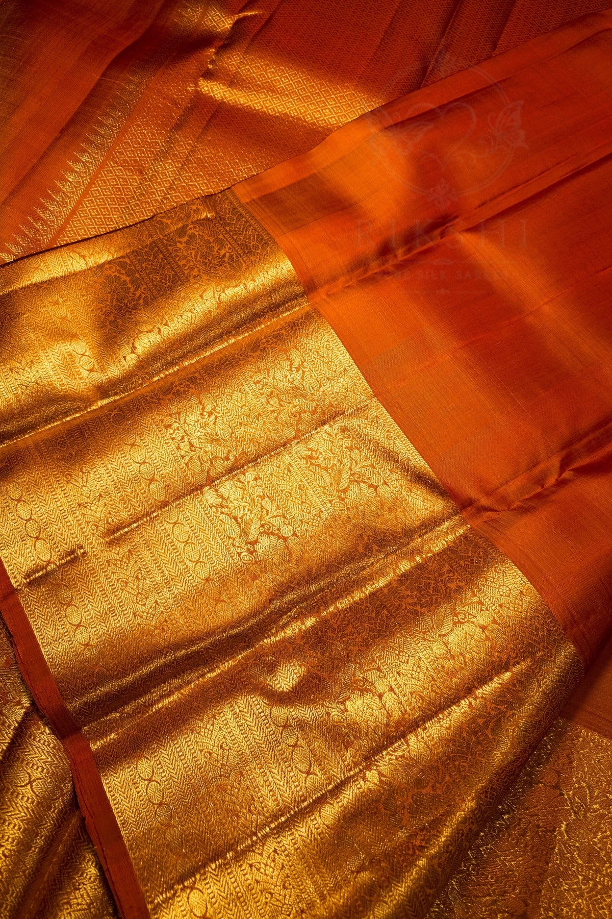 Close-up of a richly textured orange and gold fabric, likely a silk saree pallu or decorative textile, featuring intricate metallic gold brocade patterns and a solid orange border.