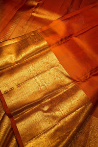 Close-up of a richly textured orange and gold fabric, likely a silk saree pallu or decorative textile, featuring intricate metallic gold brocade patterns and a solid orange border.