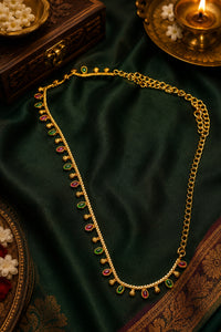 A gold-toned traditional necklace with small red and green stone accents, displayed on draped dark green satin fabric with ornate decorative props around it.