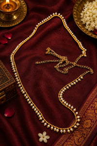 A gold-toned traditional necklace with rows of small white pearls draped elegantly on a deep red velvet fabric, styled with a brass lamp, flower petals, and decorative props.