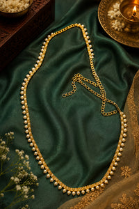 A gold chain necklace adorned with small white pearls, arranged on draped dark green satin fabric with decorative floral and brass props.