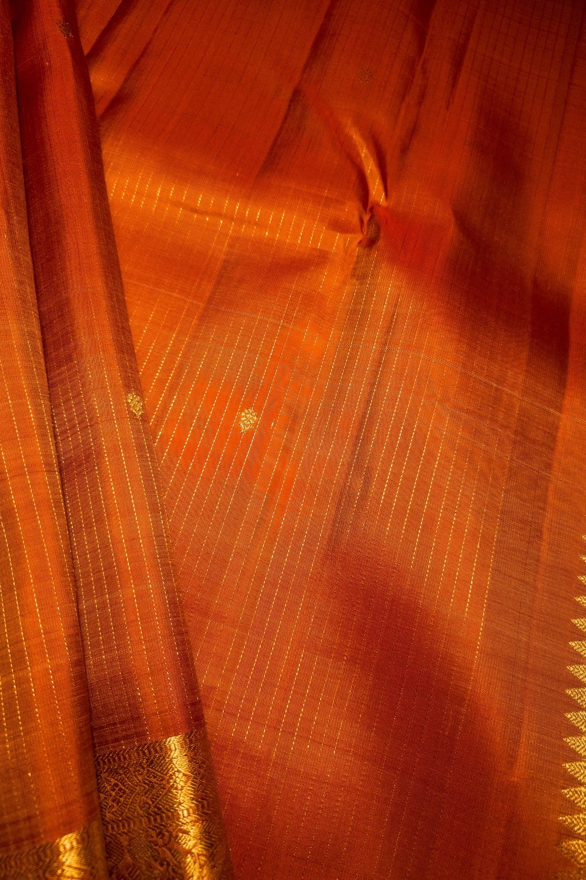 Close-up of an orange silk saree fabric with fine gold zari stripes, a gold border, and small gold motifs.