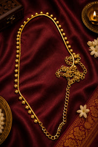 A gold-toned traditional necklace with a beaded upper section and a chain lower section, displayed on a deep red satin fabric with decorative props.