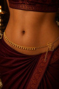 A close-up of a person's midsection wearing a gold chain belly chain (kara) with small hanging beads and a decorative side clasp, paired with a deep maroon traditional garment.