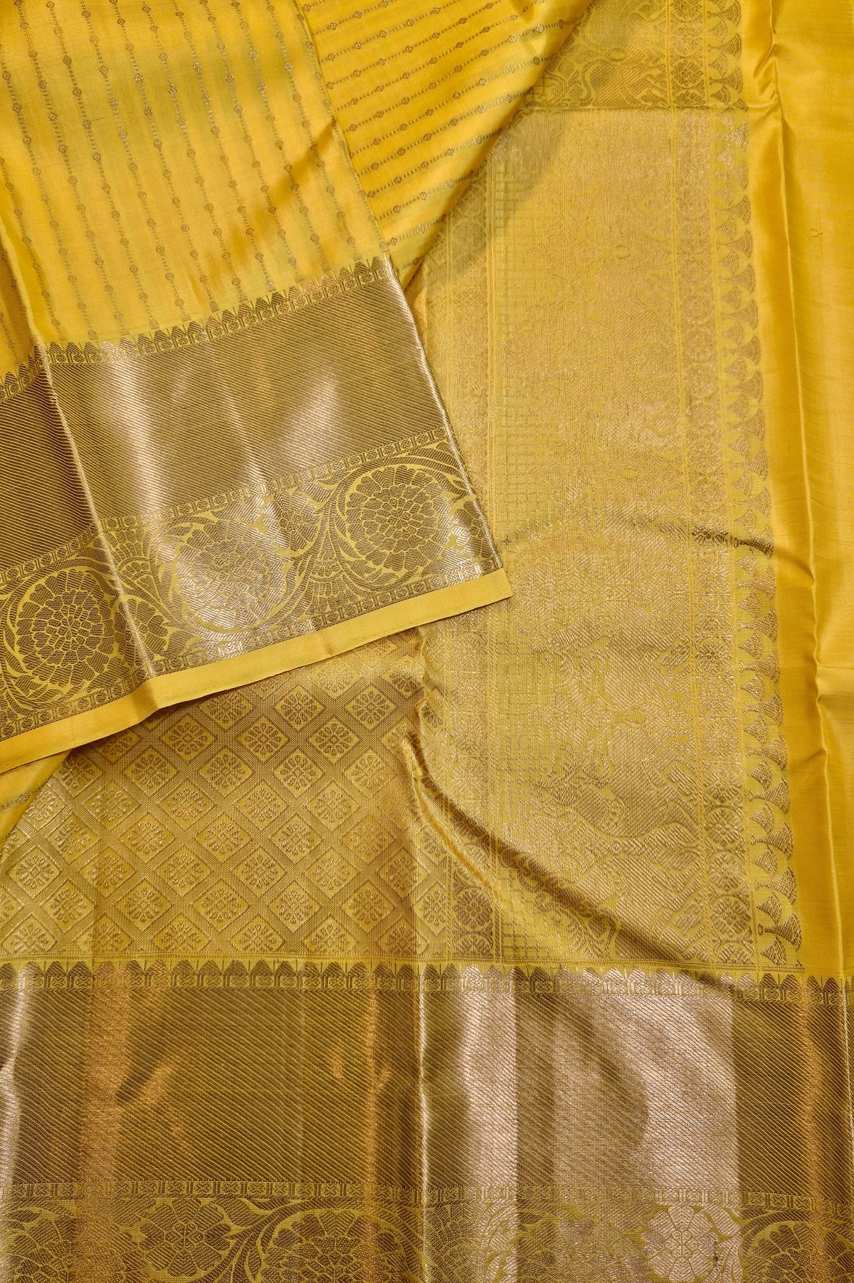 A close-up of a yellow and gold woven fabric, likely a sari, showing detailed zari embroidery, a wide gold border, and a matching yellow fabric drape.