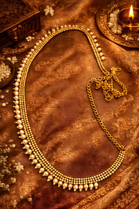 A gold-toned traditional necklace with pearl-like bead accents and a matching chain, arranged on a rich brown patterned fabric with decorative props including a lit diya and small floral elements.