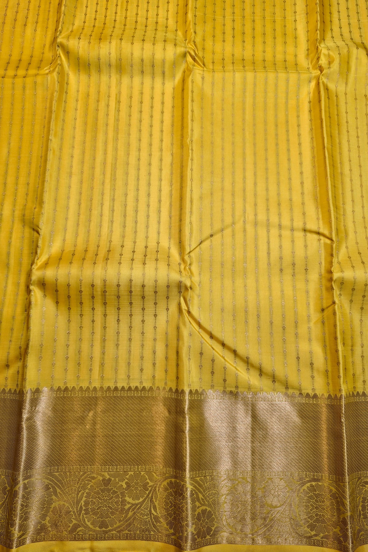 A close-up of a yellow silk saree fabric with vertical stripes and a wide metallic gold border featuring intricate floral patterns.