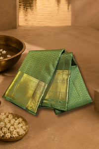 A folded green saree with a wide gold zari border and subtle vertical stripes, displayed on a warm beige surface with decorative bowls in the background.