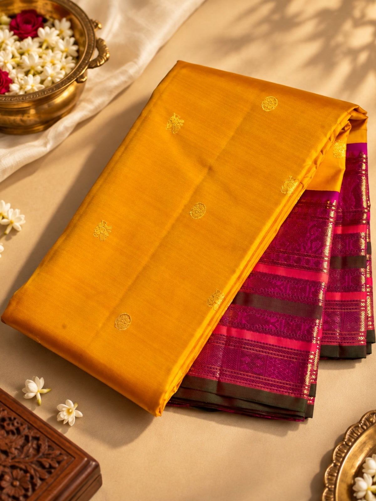 A folded yellow-gold saree with small gold motifs and a magenta-pink zari border, displayed on a beige surface with decorative floral props and a brass bowl.