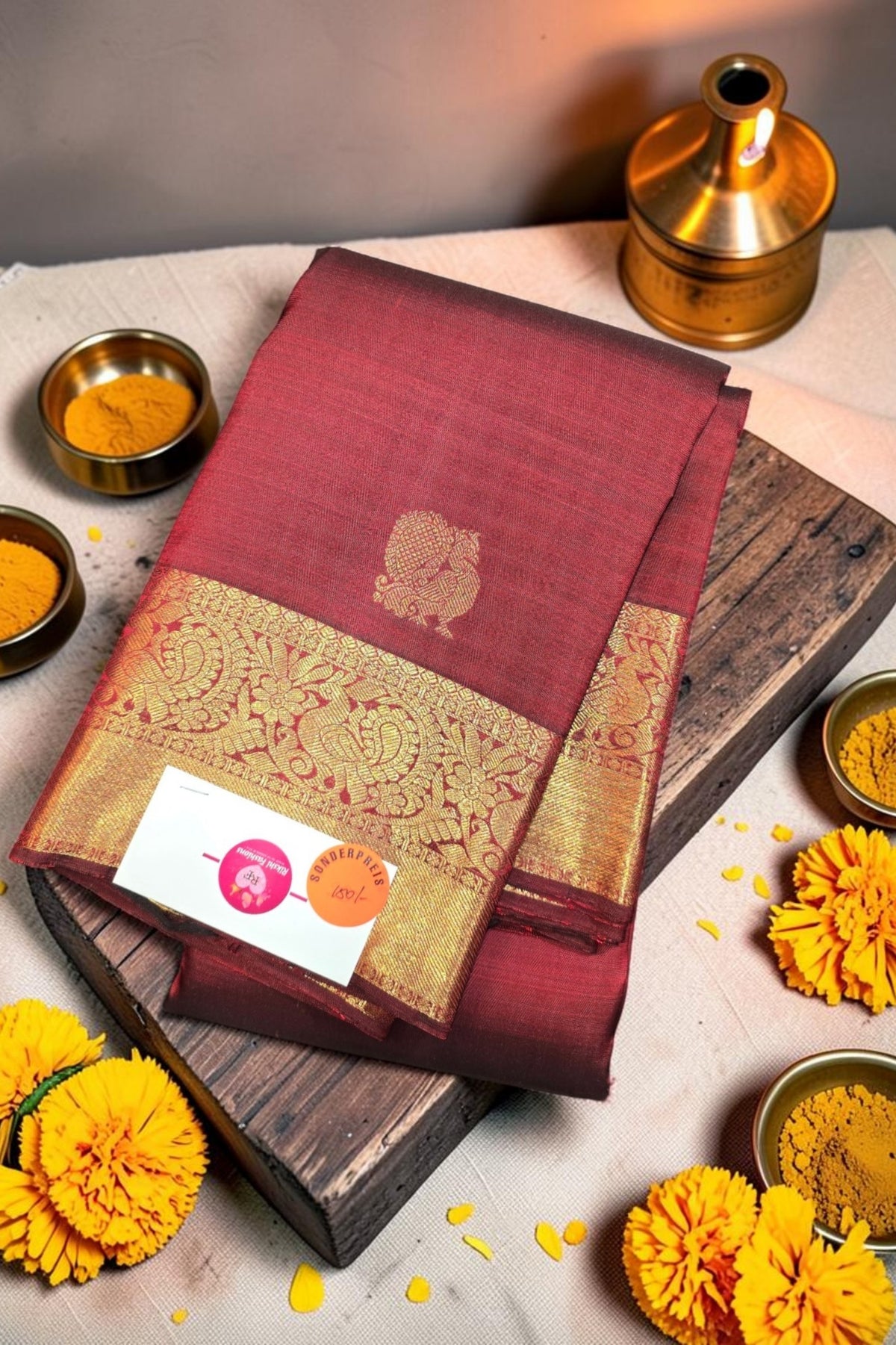 Maroon saree with gold border on a wooden board, surrounded by yellow flowers and small bowls.