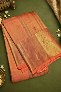 A folded traditional Indian silk saree in a warm coral-pink and gold zari weave, displayed on a dark green textured background with decorative white flowers.