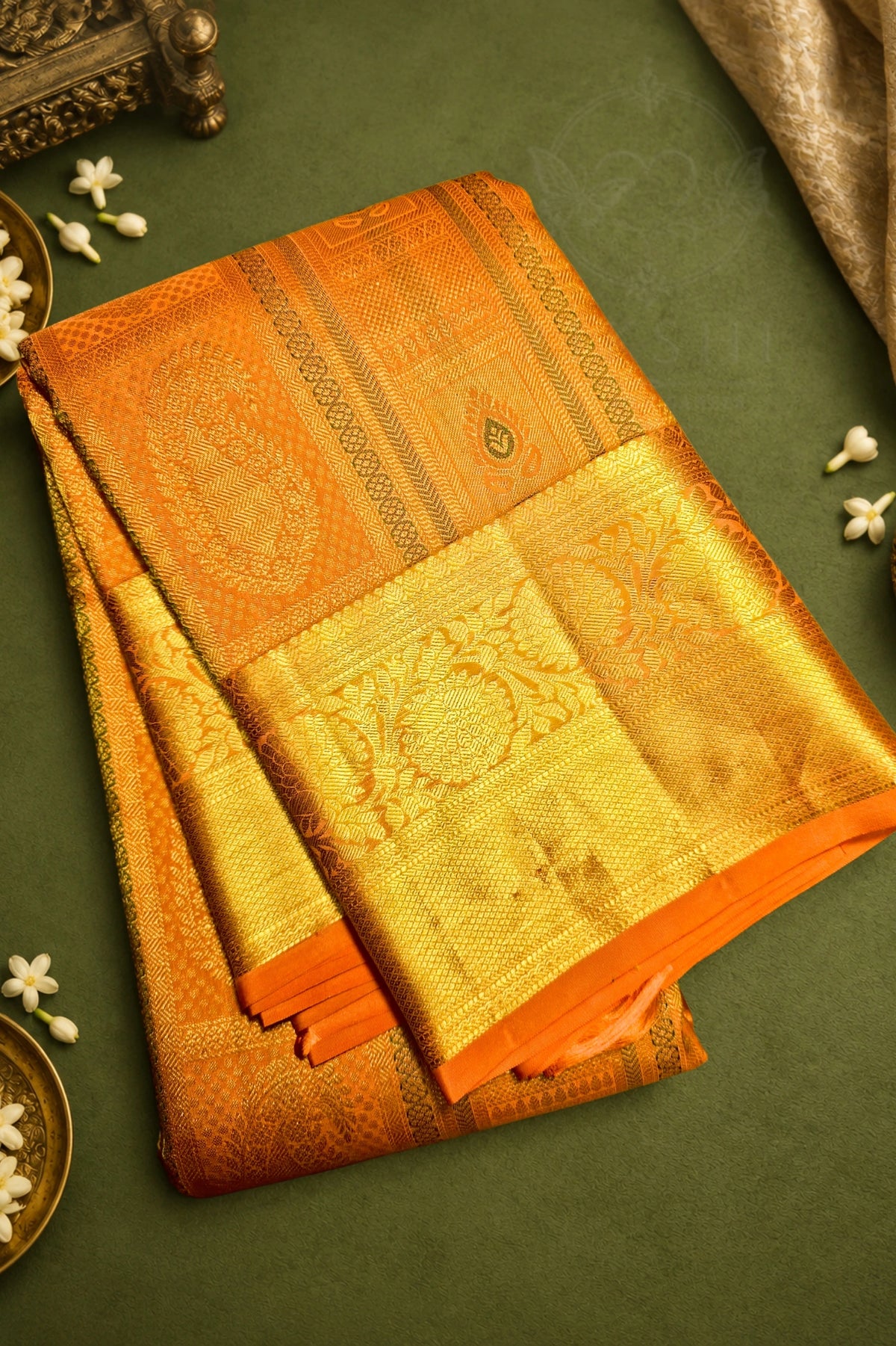 An orange and gold woven silk saree with intricate zari embroidery, neatly folded and displayed on a dark green background with decorative white flowers.