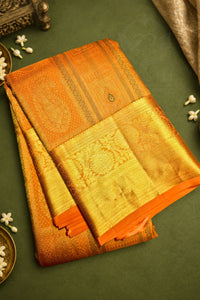 An orange and gold woven silk saree with intricate zari embroidery, neatly folded and displayed on a dark green background with decorative white flowers.