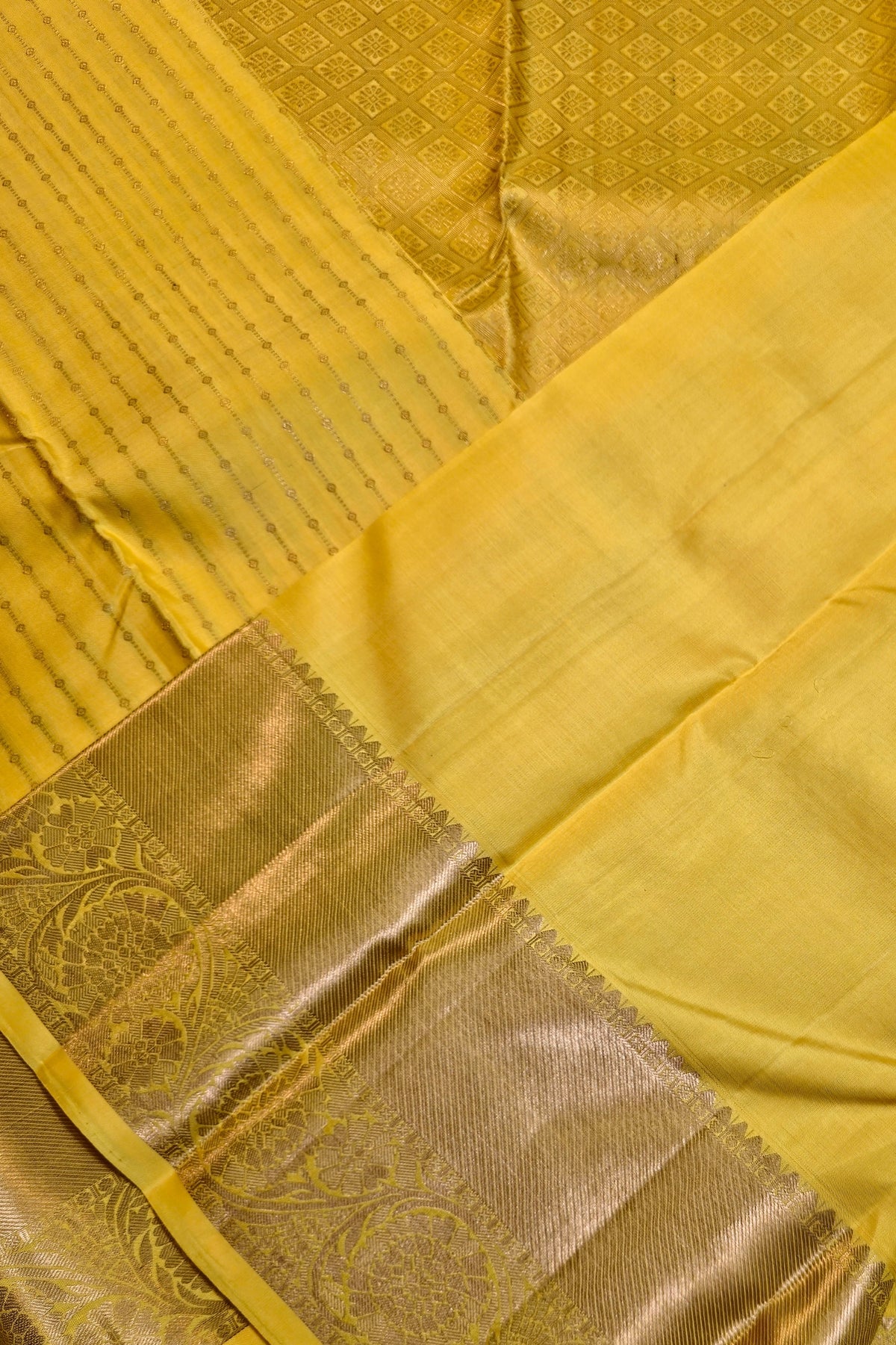 A close-up of a yellow silk saree with a wide metallic gold border featuring intricate woven floral patterns.