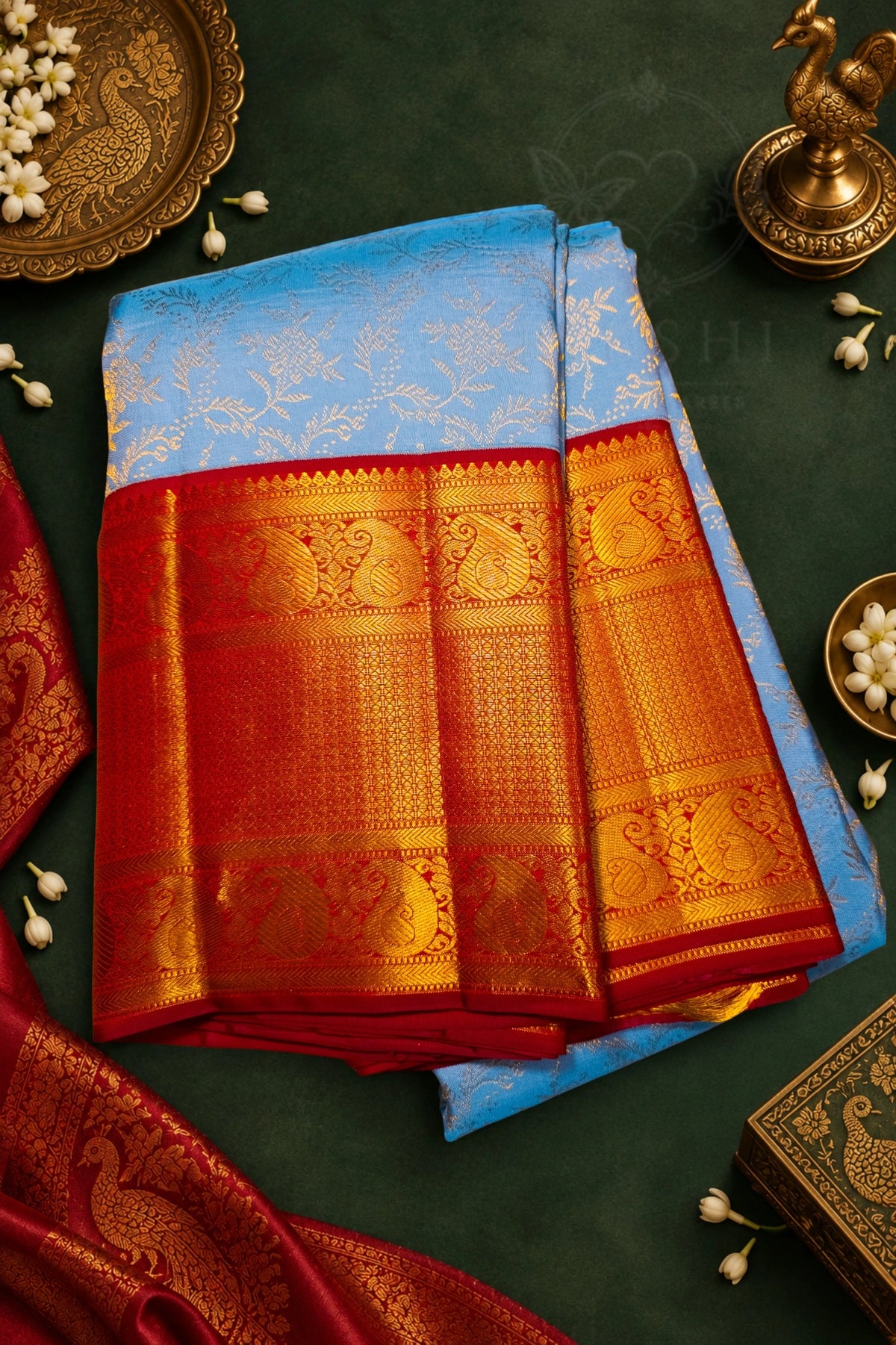 A folded blue and red silk saree with intricate gold zari embroidery, displayed on a dark green background with traditional decorative props.