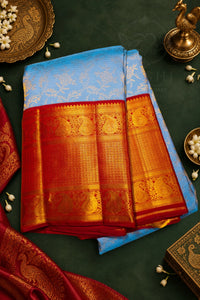 A folded blue and red silk saree with intricate gold zari embroidery, displayed on a dark green background with traditional decorative props.