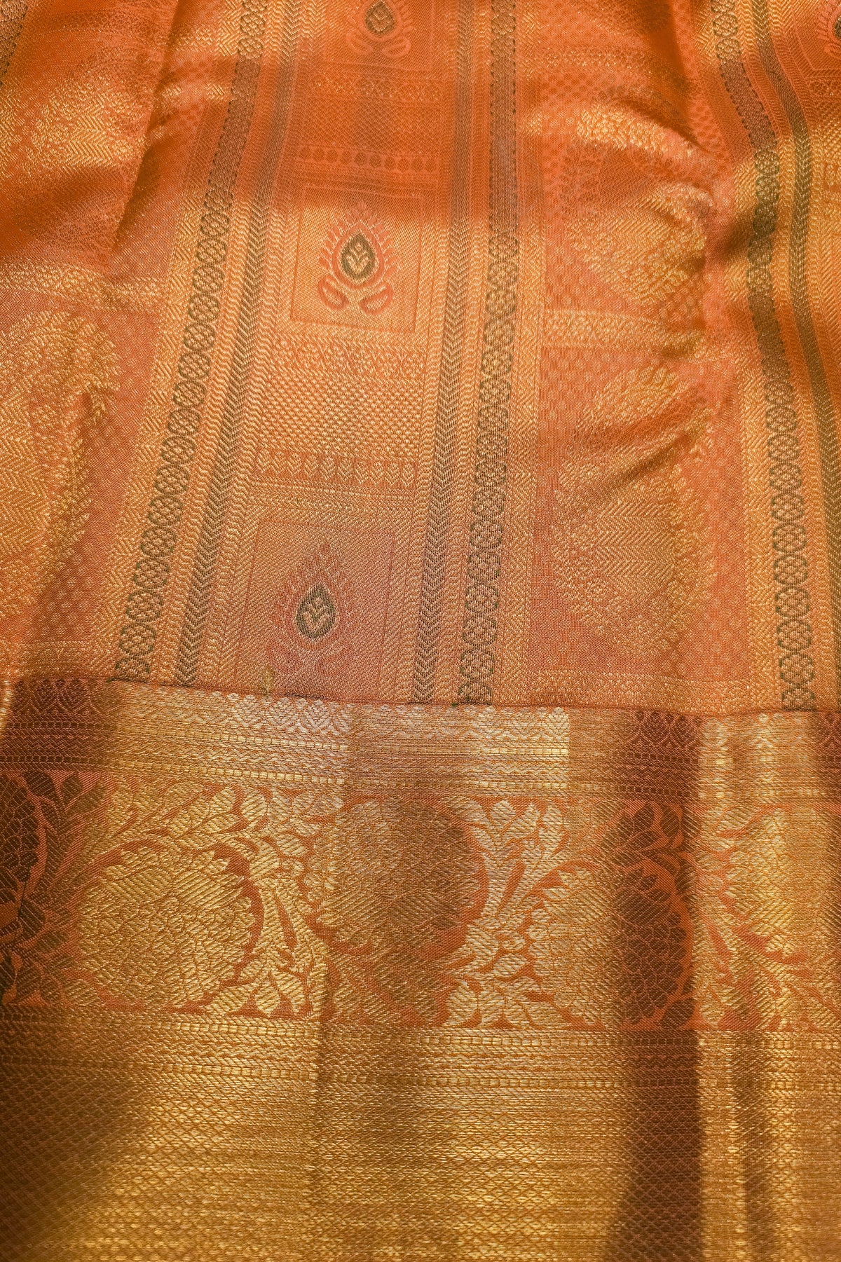 Close-up of a woven textile with a warm orange-pink base and extensive metallic gold zari patterns, including a wide decorative border.