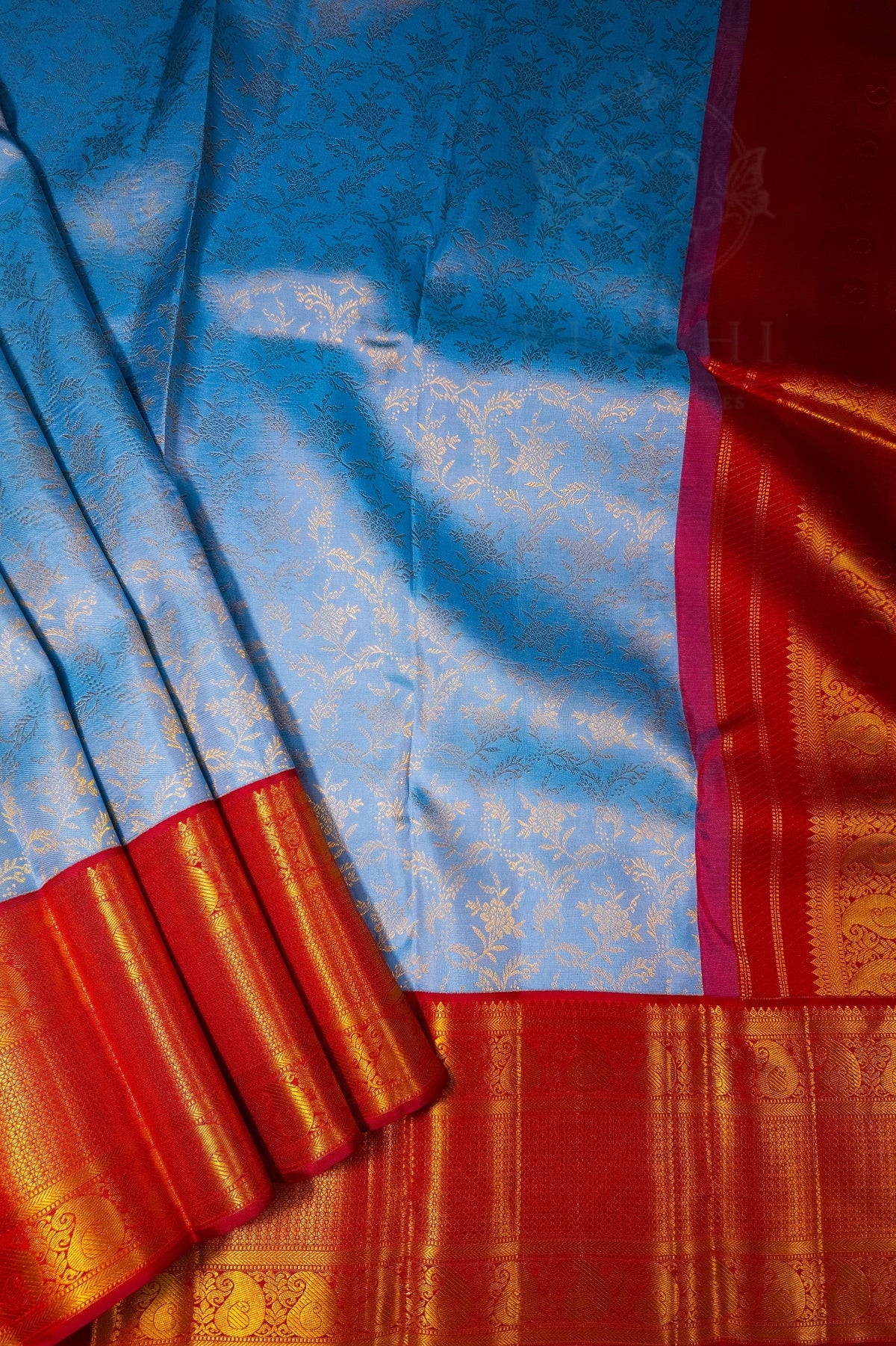 A close-up of a blue and red silk saree with gold zari brocade patterns, showing the body fabric and the decorative border.