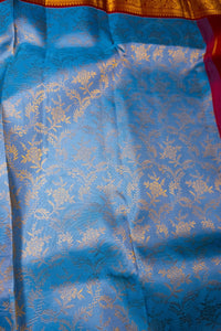 A close-up of a blue fabric with an allover gold floral brocade pattern, showing a shimmering woven texture and a glimpse of a red and gold border at the top.