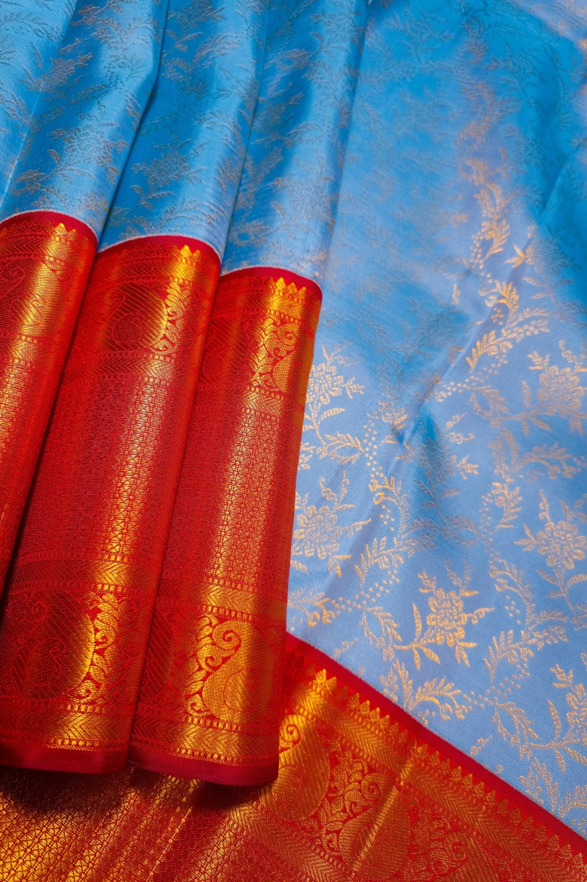 A close-up of a light blue silk saree with intricate gold floral motifs and a rich red border featuring detailed gold zari embroidery.