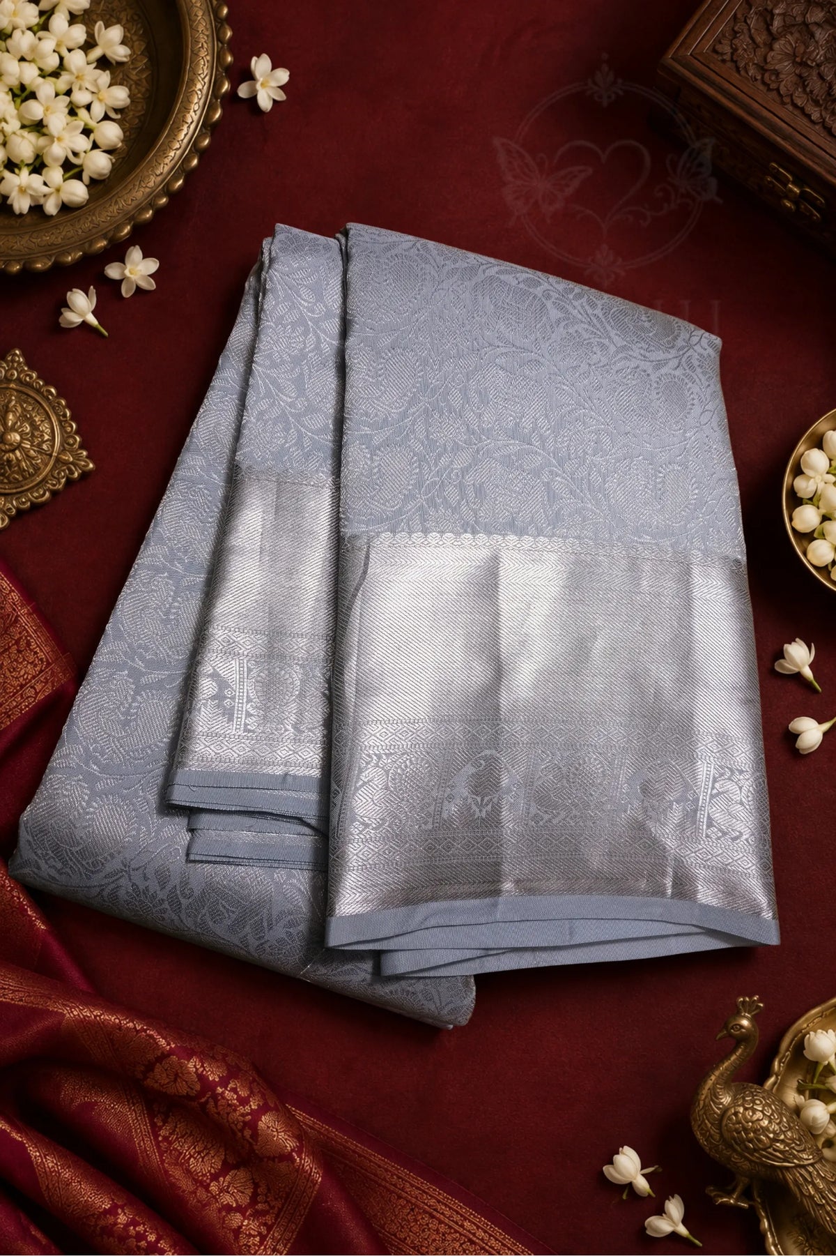A folded light blue-gray silk saree with a wide metallic silver border and subtle floral motifs, displayed on a deep red fabric background with decorative brass and white flower props.
