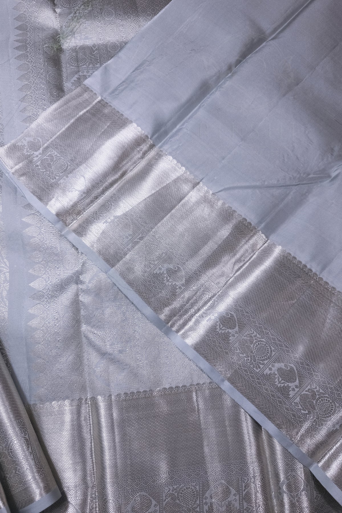 A close-up of a silver-colored silk saree with a wide, ornate brocade border and a smooth body, showing detailed woven patterns.