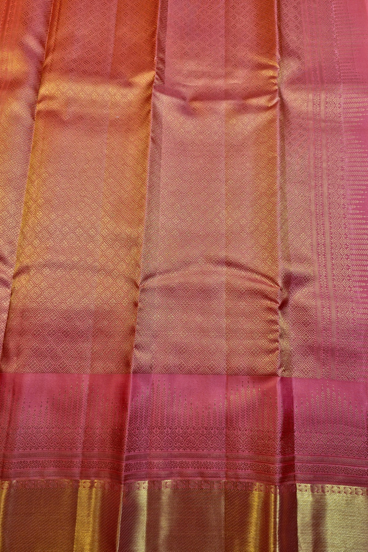 A close-up of a pink and gold zari woven fabric, likely a silk saree or textile, showing the texture, sheen, and metallic border details.