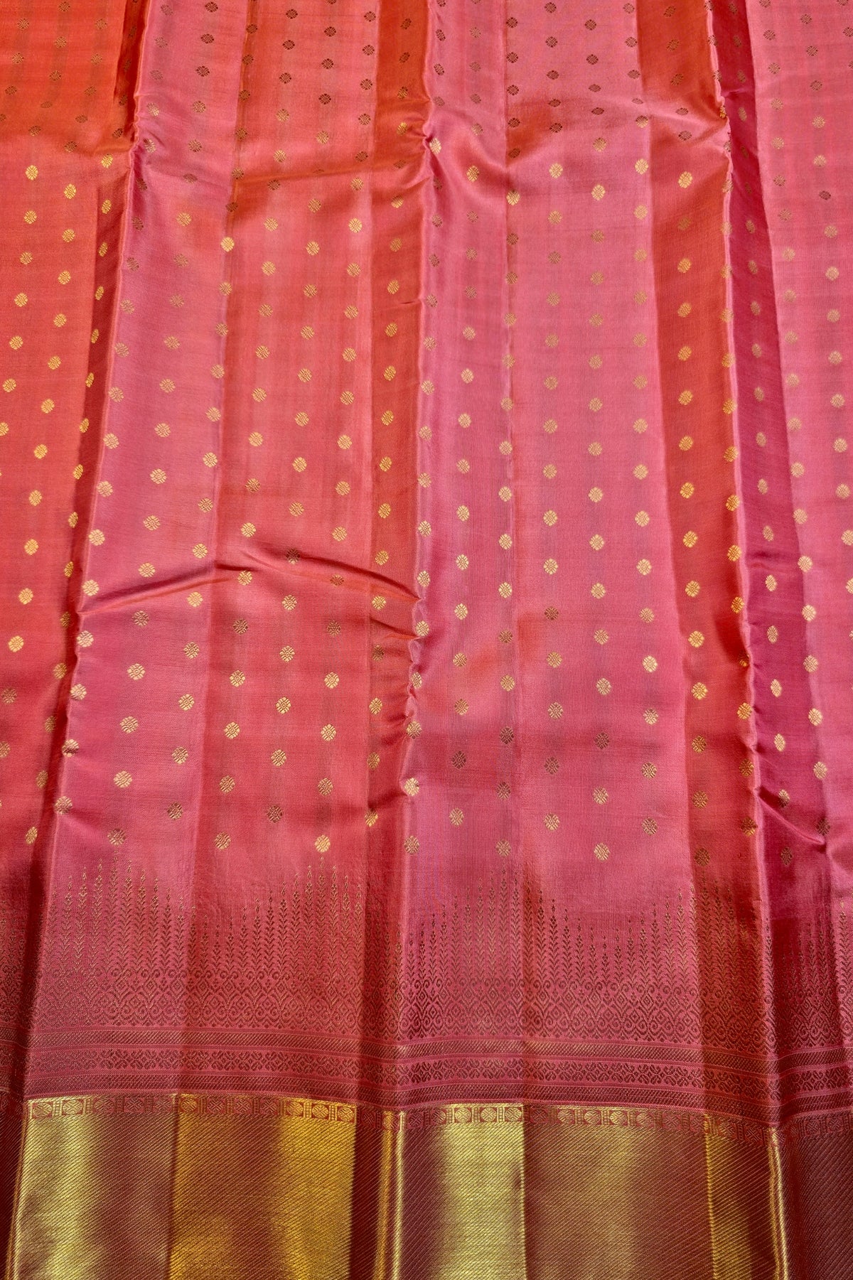 A close-up of a pink and orange-red fabric, likely a saree, decorated with small gold zari dots and a wide metallic gold border at the bottom.