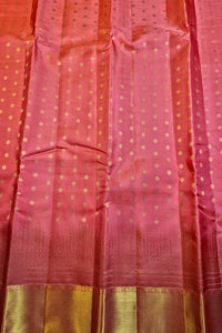 A close-up of a pink and orange-red fabric, likely a saree, decorated with small gold zari dots and a wide metallic gold border at the bottom.