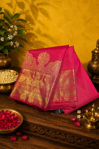 A folded bright pink silk saree with an ornate gold zari brocade pattern, displayed on a dark wooden table with traditional Indian decorative props.