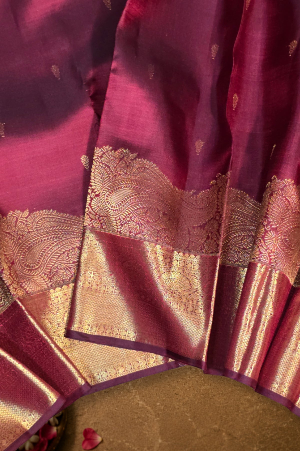 A close-up of a magenta silk saree with a wide gold zari border and intricate metallic embroidery.