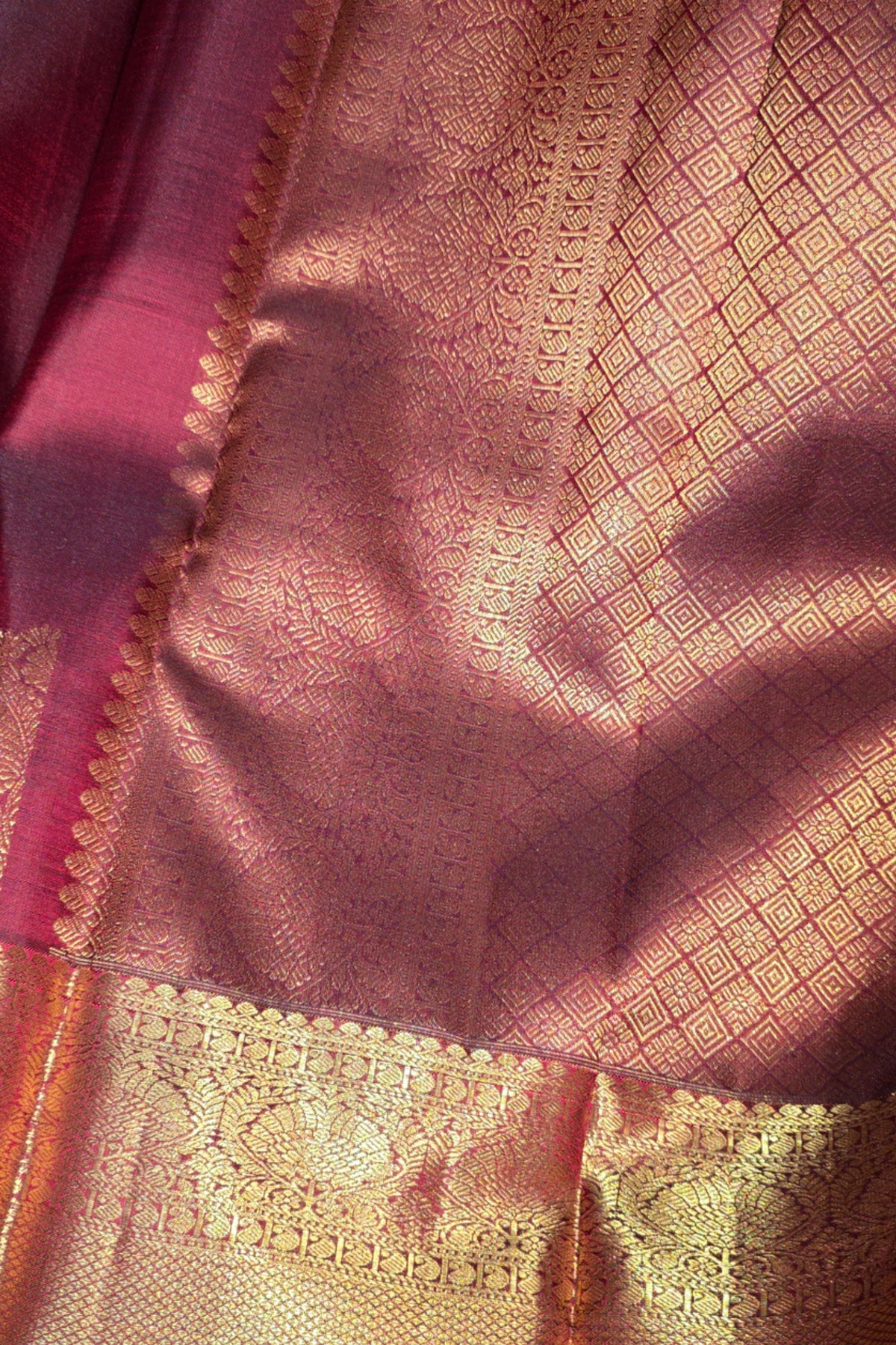 A close-up of a pink and gold woven fabric, likely a saree, featuring a rich metallic gold zari pattern, a diamond motif, and a decorative border.
