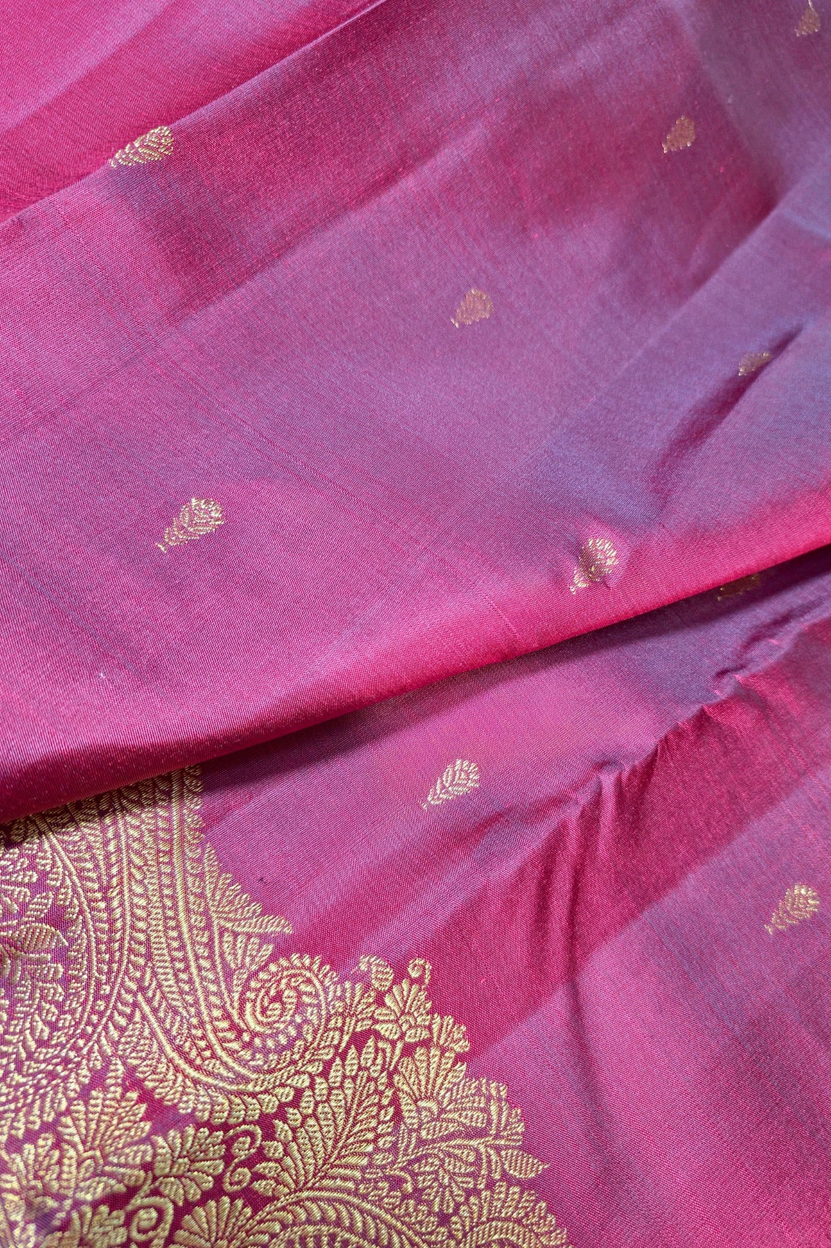 A close-up of a pink silk saree or fabric with scattered small gold motifs and a prominent gold paisley border along the edge.