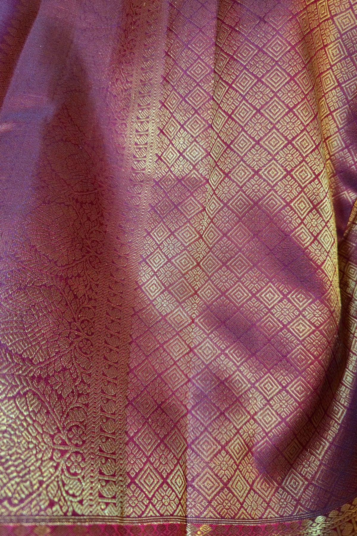 A close-up of a draped purple silk fabric with intricate gold zari woven patterns and borders.
