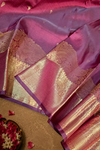 A close-up of a purple and pink silk saree with intricate gold zari woven borders, draped elegantly on a neutral surface.