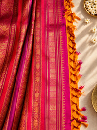 A close-up of a richly patterned sari with a magenta-pink body, gold zari woven motifs, and a contrasting orange border with tassel fringe, styled on a light neutral surface with small white flower decorations.