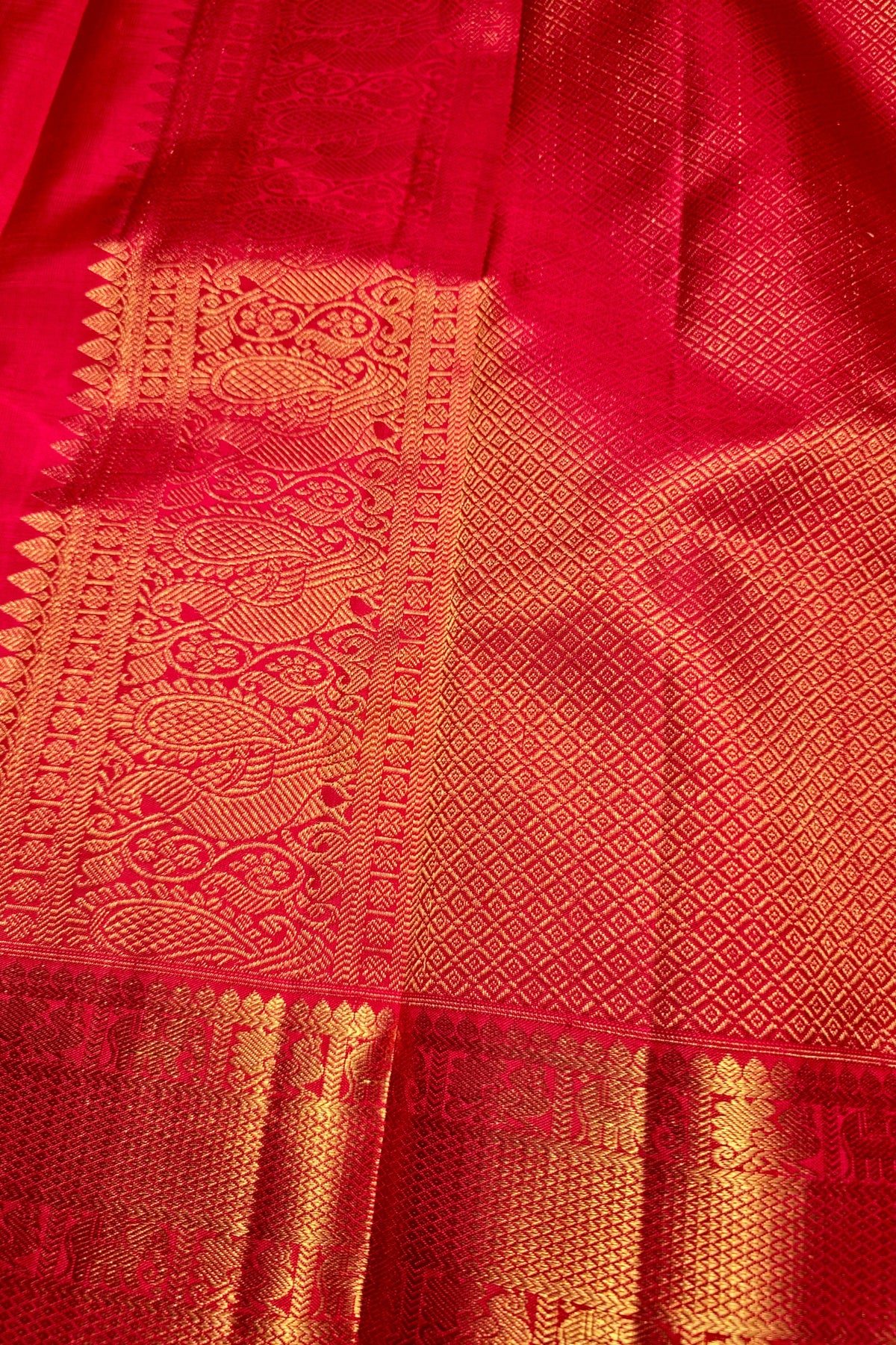 A close-up of a rich red fabric with intricate gold zari woven patterns and borders, likely a silk saree or textile.