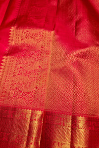 A close-up of a rich red fabric with intricate gold zari woven patterns and borders, likely a silk saree or textile.