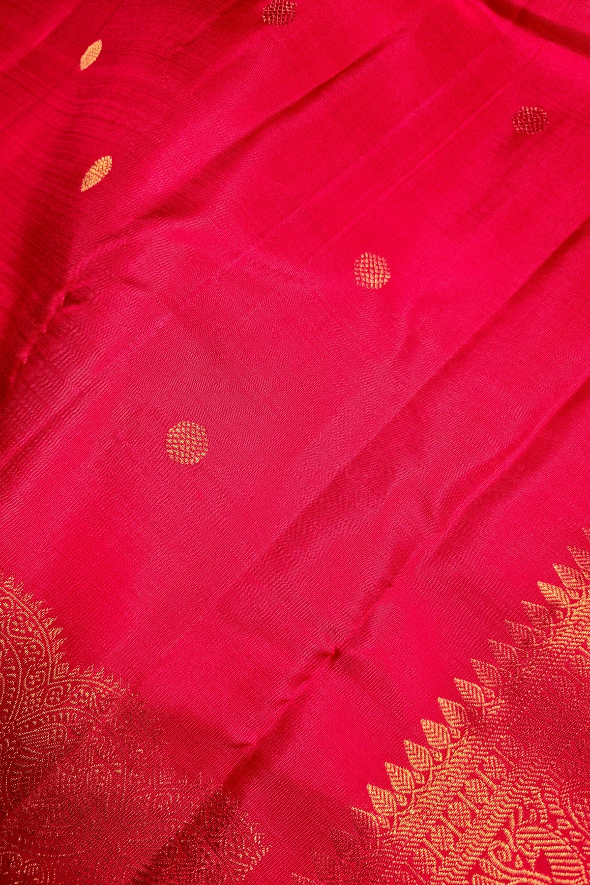 A close-up of a bright pink silk fabric, likely a Banarasi silk saree, featuring small gold zari woven dots and a decorative gold border pattern along the edge.