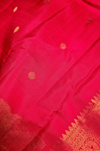 A close-up of a bright pink silk fabric, likely a Banarasi silk saree, featuring small gold zari woven dots and a decorative gold border pattern along the edge.