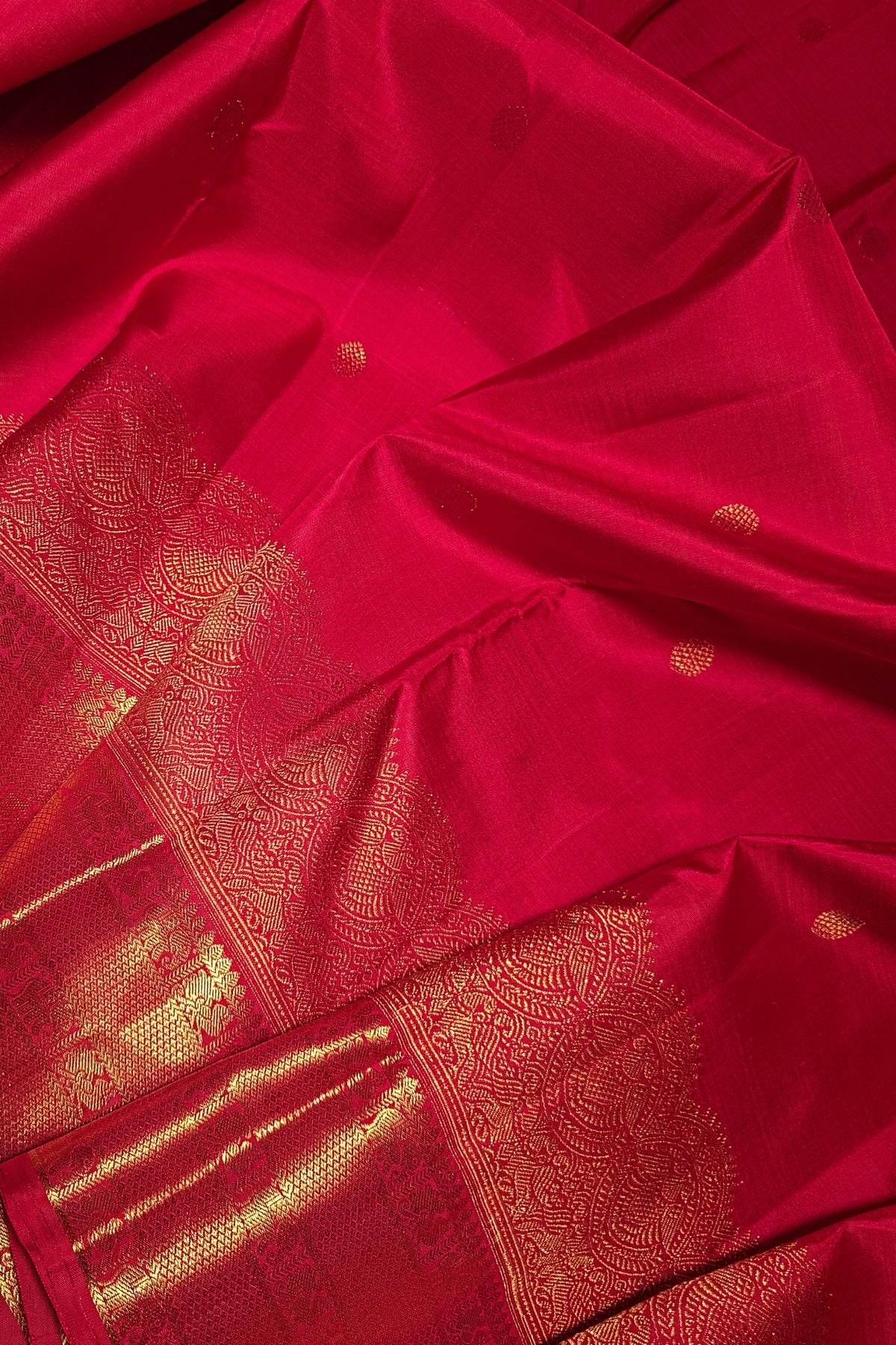 A close-up of a rich red silk saree with intricate gold zari brocade work and small gold motifs scattered across the fabric.
