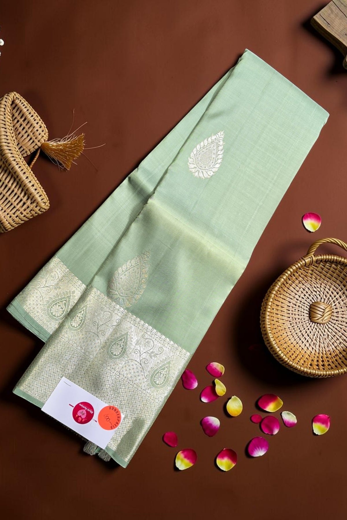 Green saree with white patterns on a brown surface with a woven basket and rose petals.