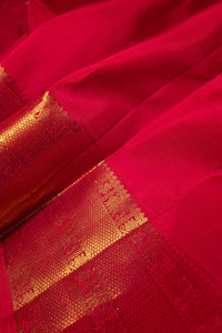 A close-up of a bright red fabric, likely a saree, featuring a wide gold zari brocade border with traditional motifs.
