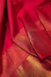 A close-up of a rich red silk fabric, likely a Banarasi sari, featuring intricate gold zari woven patterns and borders.