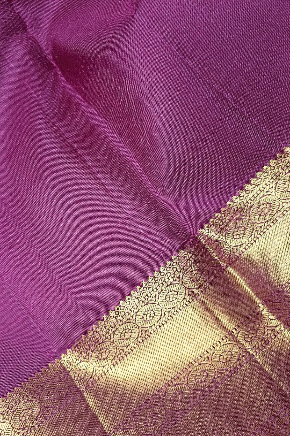 A close-up of a purple silk fabric, likely a saree, featuring a rich metallic gold zari border with intricate woven patterns.