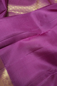 A close-up of a purple silk saree fabric with a gold zari woven border pattern visible in the upper left and lower left corners.